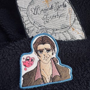 May include: Embroidered patches featuring a cartoon illustration of a man with brown hair, wearing a brown jacket and pink shirt, with a pink blob. A second patch has the text "Magical Little Stitches" in a circle.
