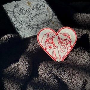 May include: Red heart-shaped embroidered patch with a white background featuring a cartoon couple. The couple is holding hands and smiling. The text "Magical Little Stitches" is visible in the background.