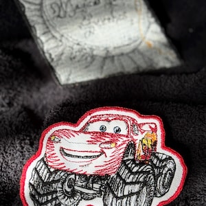 May include: Red monster truck with a smiling face, black wheels, and a red outline. The truck is embroidered on a black fabric.
