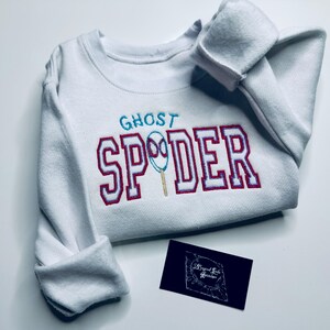 May include: White sweatshirt with the text "GHOST SPIDER" embroidered in teal and red. The design includes a spider web graphic. A small black business card is in the lower right corner.