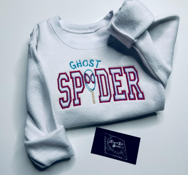 May include: White sweatshirt with the text "GHOST SPIDER" embroidered in teal and red. The design includes a spider web graphic. A small black business card is in the lower right corner.
