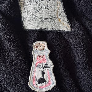 May include: A pink and white embroidered patch of a potion bottle with a black silhouette of a witch inside. The patch has a white border and a brown cork on top. The text "Magical Spells Stitches" is embroidered on a separate patch.