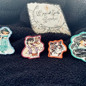 May include: Embroidered patches featuring characters from a popular animated film. The patches include a princess in a teal dress, a character with a monkey, a tiger, and another princess. A rectangular patch with the text "Magical Little Stitches" is also present.