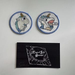 May include: Two round embroidered patches featuring a cartoon unicorn with blue and white details. One unicorn has its mouth open. A black card with the text "Magical Little Stitches" is below.