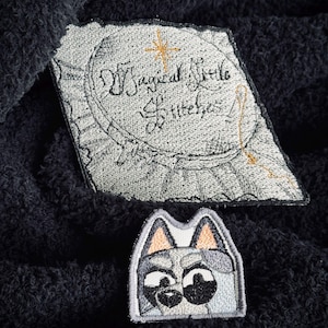 May include: Two embroidered patches. The first patch is gray with white stitching and the text "Magical Style Stitches". The second patch is gray with white stitching and depicts a cartoon dog's face.