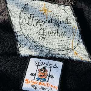 May include: Two embroidered patches on a black background. One patch reads "Magical Little Stitches" with a gold star. The other patch says "Wanted" with a cartoon character and "$50 Billion Reward".