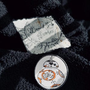 May include: A white and orange embroidered patch of a Star Wars BB-8 droid character on a black fabric background.