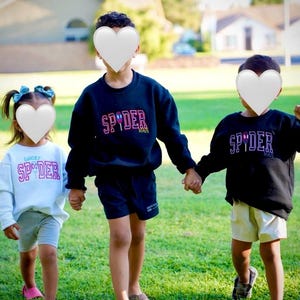 May include: Three children wearing matching sweatshirts and shorts stand on a grassy lawn. The sweatshirts are black and white, with the word "SPYDER" in a colorful font. The children are holding hands.