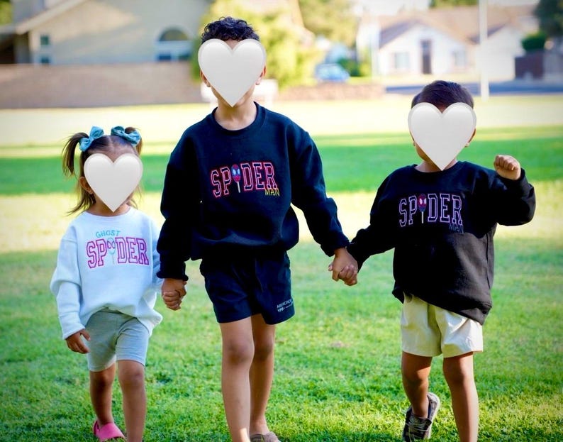 May include: Three children wearing matching sweatshirts and shorts stand on a grassy lawn. The sweatshirts are black and white, with the word "SPYDER" in a colorful font. The children are holding hands.