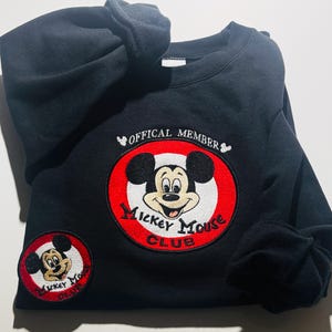 May include: Black sweatshirt featuring a large embroidered Mickey Mouse Club patch with a red and white circular design. The sweatshirt also has a smaller matching patch. The text "OFFICAL MEMBER" is embroidered above the large patch.