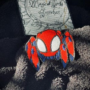 May include: A red and blue embroidered spider patch with white eyes. The patch is on a dark gray fuzzy surface. The text "Magical Little Stitches" is visible on a white background.