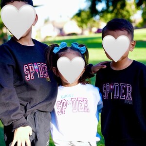 May include: Three children wearing black and white sweatshirts with the word "SPIDER" in a red and white design. The child in the middle is wearing a white shirt with the text "GHOST SPIDER". The background is a green grassy area.