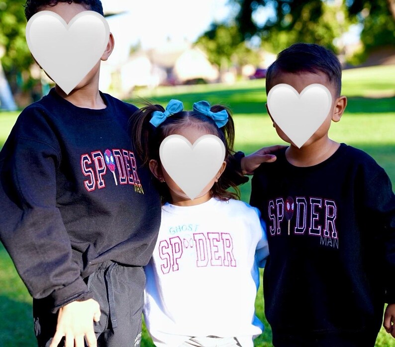 May include: Three children wearing black and white sweatshirts with the word "SPIDER" in a red and white design. The child in the middle is wearing a white shirt with the text "GHOST SPIDER". The background is a green grassy area.