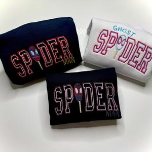 May include: Three embroidered sweatshirts. One navy blue with "SPIDER MAN" in red and white lettering, and a spider-themed popsicle graphic. Another is black with the same design. The third is white with "GHOST SPIDER" in pink lettering and a ghost-themed popsicle graphic.