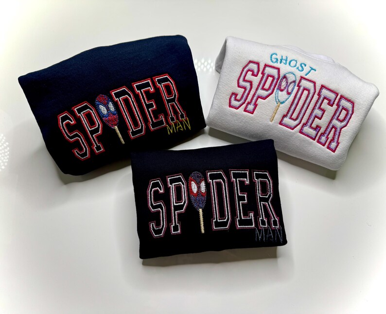 May include: Three embroidered sweatshirts. One navy blue with "SPIDER MAN" in red and white lettering, and a spider-themed popsicle graphic. Another is black with the same design. The third is white with "GHOST SPIDER" in pink lettering and a ghost-themed popsicle graphic.