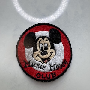 May include: Embroidered patch featuring Mickey Mouse with a smiling face, black ears, and a red and white circular border. The text "Mickey Mouse Club" is embroidered in black on the bottom of the patch.