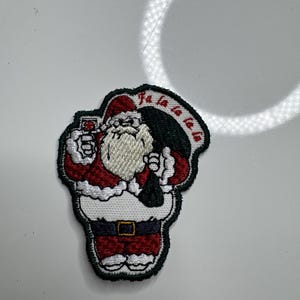 May include: Embroidered patch featuring Santa Claus in a red suit, holding a glass of wine and a sack. The patch has a white beard and the words "Fa la la la la" in red. The patch is outlined in green.