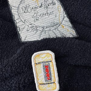 May include: Two embroidered patches. The first patch is white with black stitching and features a circle with the text "Magical Stitch" and "Stitches" inside. The second patch is yellow with red and black stitching and features a stylized image of a can of soda.