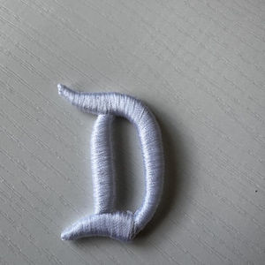 May include: A white embroidered letter "D" patch. The patch is made of tightly woven threads, creating a textured appearance. The letter has a classic, rounded serif design, suitable for various crafting projects.