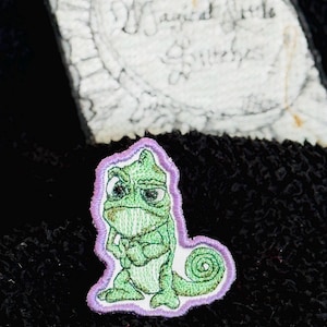 May include: Embroidered patch featuring a green chameleon with a grumpy expression, outlined in purple. The patch is on a black textured surface. In the background, a white tag with the text "Magical Little Giggles" is visible.