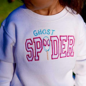 May include: White long-sleeved sweatshirt with the text "GHOST SPDER" embroidered in teal and pink. The design includes a ghost-shaped spider illustration. The sweatshirt is a children's apparel item.