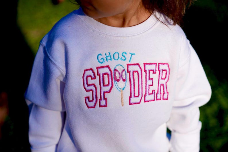May include: White long-sleeved sweatshirt with the text "GHOST SPDER" embroidered in teal and pink. The design includes a ghost-shaped spider illustration. The sweatshirt is a children's apparel item.