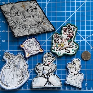 May include: A set of embroidered iron-on patches featuring characters from Disney's Cinderella. The patches include Cinderella in her ball gown, Cinderella in her everyday dress, a pillow, two mice, and a sign that says "Magical Bottle of Wishes".