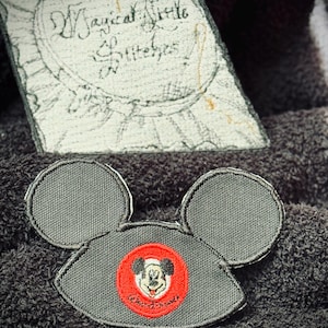 May include: A black and gray Mickey Mouse ear patch with a red circle featuring a cartoon image of Mickey Mouse. The circle has the text "Mickey Mouse" in white.