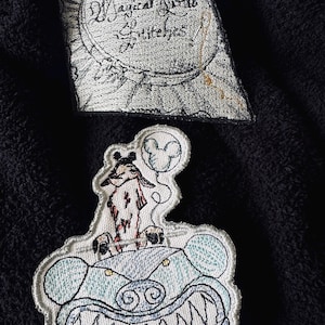 May include: Two embroidered patches. The first patch is white with black stitching and features the text "Magical Little Stitches". The second patch is white with black and blue stitching and features a cartoon character driving a blue car with a Mickey Mouse balloon.