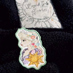 May include: Embroidered patch featuring a princess with long blonde hair adorned with flowers, a sun emblem, and a green border. The patch is on a black surface, with a second patch in the background that reads "Magical Little Stitches."