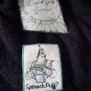 May include: Two embroidered patches. The first patch is white with black embroidery and says "Magical Little Stitchers". The second patch is white with colorful embroidery and shows a cartoon character holding a plate of food. The text below the character says "Spinach Puff?"