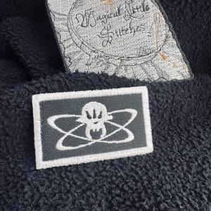 May include: Two embroidered patches on a dark blue textured surface. The top patch is a light gray irregular shape with the words "Magical Little Stitches" in black script. The bottom patch is rectangular, dark gray with a white skull and atom design.