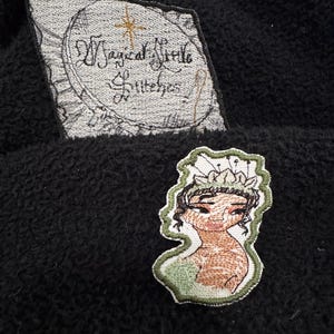 May include: Black fabric with two embroidered patches. One patch features the text "Magical Little Stitches" in a circular design. The other patch is an illustration of a woman wearing a crown and a green dress.