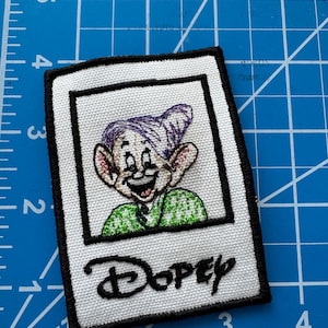 May include: A white embroidered patch with a picture of Dopey from Snow White and the Seven Dwarfs. Dopey is wearing a green shirt and a purple hat. The patch has a black border and the word "Dopey" is embroidered in black below the picture.