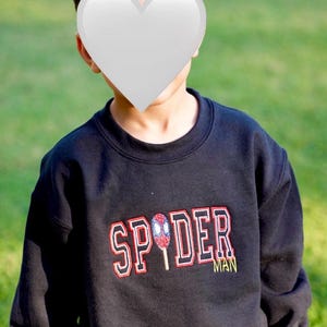 May include: Black sweatshirt with the word "SPIDER" in red and white lettering, and a Spider-Man graphic. The word "MAN" is in yellow. The sweatshirt is worn by a person.