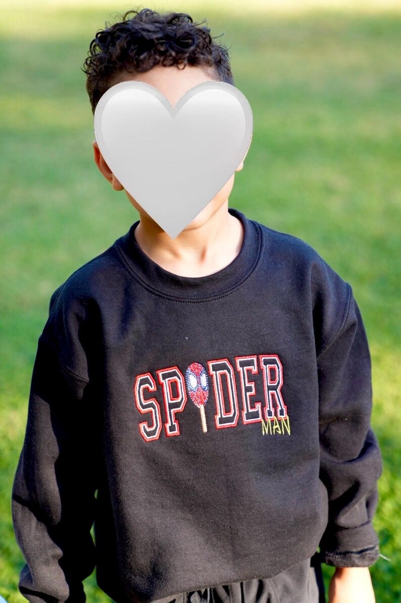 May include: Black sweatshirt with the word "SPIDER" in red and white lettering, and a Spider-Man graphic. The word "MAN" is in yellow. The sweatshirt is worn by a person.