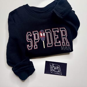 May include: Black sweatshirt with the word "SPIDER" in white and red lettering, and the word "MAN" embroidered below. A Spider-Man-themed design is embroidered in the center. A small, dark blue business card is in the lower right corner.