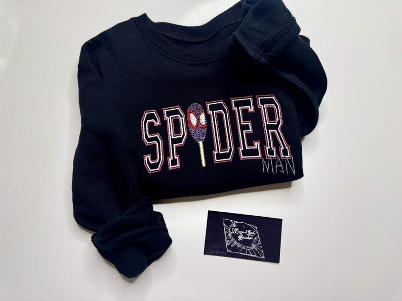 May include: Black sweatshirt with the word "SPIDER" in white and red lettering, and the word "MAN" embroidered below. A Spider-Man-themed design is embroidered in the center. A small, dark blue business card is in the lower right corner.