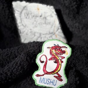 May include: Red and yellow embroidered dragon cartoon character patch with the word "Mushu" in green lettering on a white background.