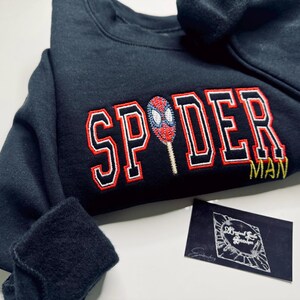 May include: A navy blue sweatshirt with the word "SPIDER" in red and white block letters, and a Spiderman graphic. The word "MAN" is below in yellow. A small black card is in the bottom right corner.