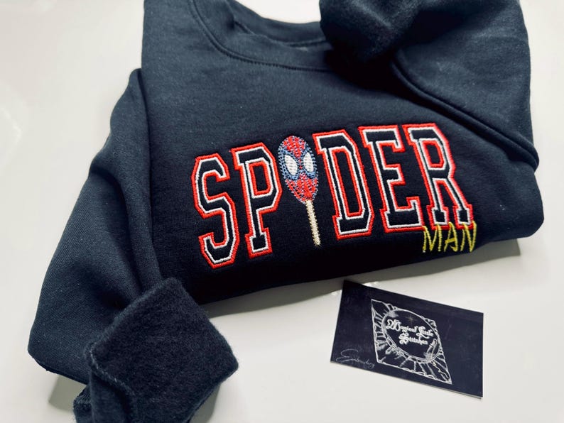 May include: A navy blue sweatshirt with the word "SPIDER" in red and white block letters, and a Spiderman graphic. The word "MAN" is below in yellow. A small black card is in the bottom right corner.