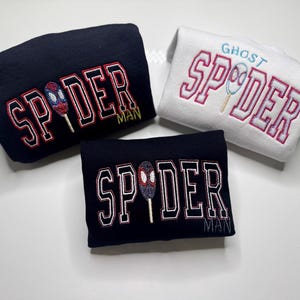 May include: Three folded sweatshirts. Two are navy blue with "SPIDER MAN" in red and white lettering, and a spider-themed graphic. The third is white with "GHOST SPIDER" in pink lettering and a spider graphic.
