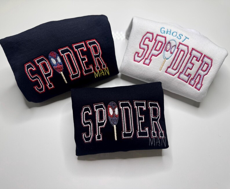May include: Three folded sweatshirts. Two are navy blue with "SPIDER MAN" in red and white lettering, and a spider-themed graphic. The third is white with "GHOST SPIDER" in pink lettering and a spider graphic.