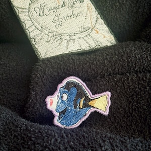 May include: Embroidered patch featuring a blue fish with a yellow fin and a pink mouth. The patch is on a dark, textured fabric. A second patch with the text "Magical Little Stitches" is also visible.
