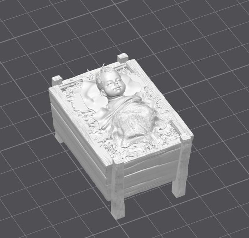 Op de afbeelding: Grijswaarden sculptuur van een baby in een houten kribbe. De baby is in een deken gewikkeld en rust op een kussen. De kribbe heeft een stro-achtig materiaal binnenin. De sc&egrave;ne is een weergave van een kerststal.