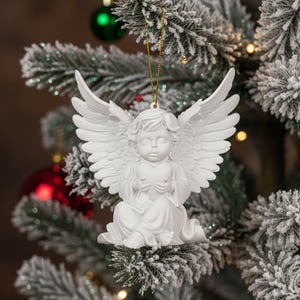 Op de afbeelding: Een witte kerstversiering in de vorm van een engel met uitgespreide vleugels en een hart, hangend aan een besneeuwde kerstboom. De engel is gemaakt van een glad materiaal en heeft een gouden touwtje om op te hangen. Rode en groene ballen zijn zichtbaar op de achtergrond.