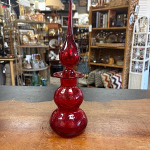 May include: A vibrant red glass decanter featuring a bulbous, double-tiered base and a long, slender, pointed stopper. The glass has a subtle crackled texture throughout, and the piece stands upright with a distinct, elongated silhouette.
