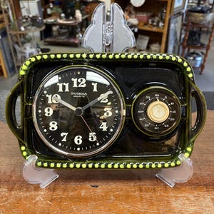 May include: A rectangular dark green ceramic electric clock with a black face, white numerals, and the text Dugena electric. It features a secondary circular timer dial on the right side and a decorative dotted border along the outer edge.