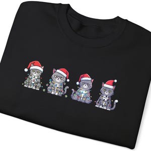 Peut inclure: Sweat-shirt noir avec quatre chats gris grincheux portant des bonnets de Père Noël et des guirlandes lumineuses. Les chats ont une expression renfrognée. Un design amusant et festif pour les fêtes de fin d'année.