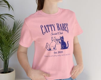 May include: Pink t-shirt with a graphic of two cats toasting with glasses. The text reads "Catty Babes Social Club Est. 2025 Where the purrfectly sassy meet to spill the tea."